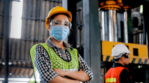 Female worker in a factory looking straight into the camera, crossed arms, wearing a safety vest, mask, goggles and helmet